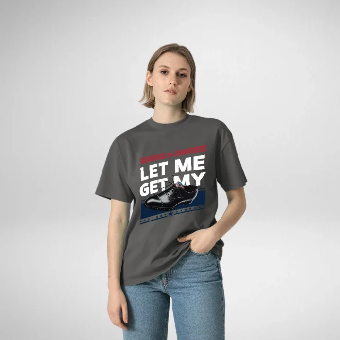 Woman posing in a gray t-shirt featuring the phrase 'LET ME GET MY' alongside an illustration of a polished black shoe, with her hand casually in the pocket of her jeans.